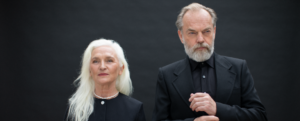 Actors Olwen Fouere and Hugo Weaving stand in black clothing against a grey backdrop, looking stern.